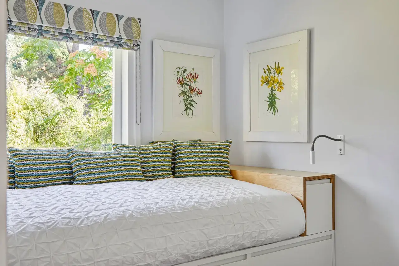 Cama en habitación de 6 metros cuadrados, con cojines coloridos, cuadros de flores y ventana con vistas al jardín.