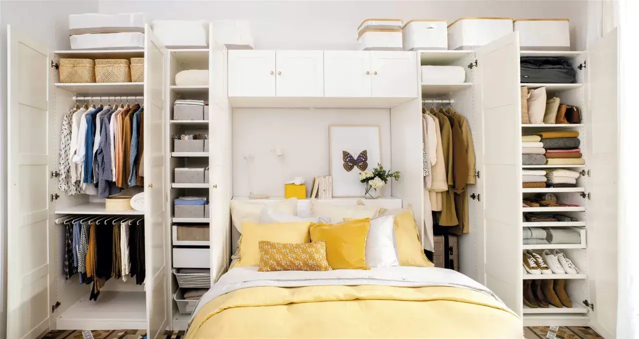 Cama con almohadas amarillas y blancas, rodeada de armarios blancos llenos de ropa y cestas. El espacio entre cama y armario está bien aprovechado.