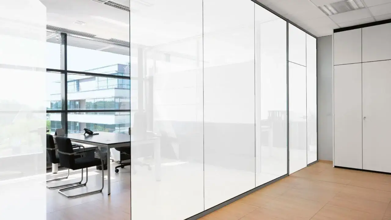 Oficina moderna con paredes de cristal inteligente que reflejan la luz. Se ven mesas y sillas, y un edificio exterior.