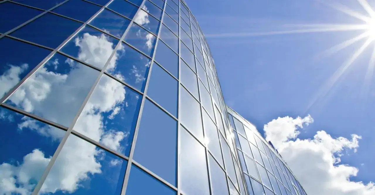 Edificio moderno con fachada de vidrio reflectante que muestra el cielo azul y nubes. El sol brilla intensamente, destacando el factor solar del vidrio.