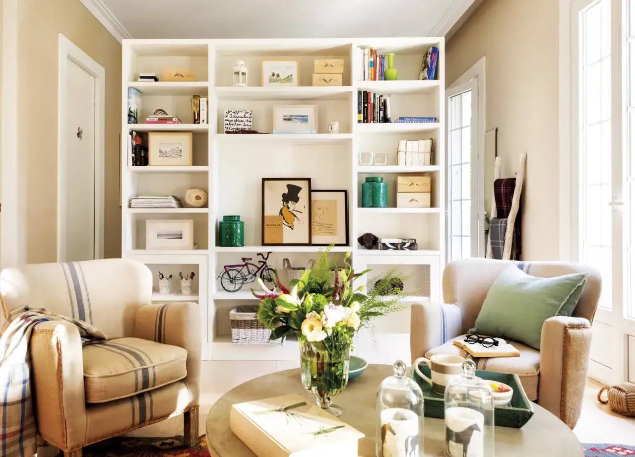 Dos sillones c&oacute;modos frente a una estanter&iacute;a blanca llena de libros y objetos decorativos, creando un rinc&oacute;n acogedor en estas casas peque&ntilde;as.