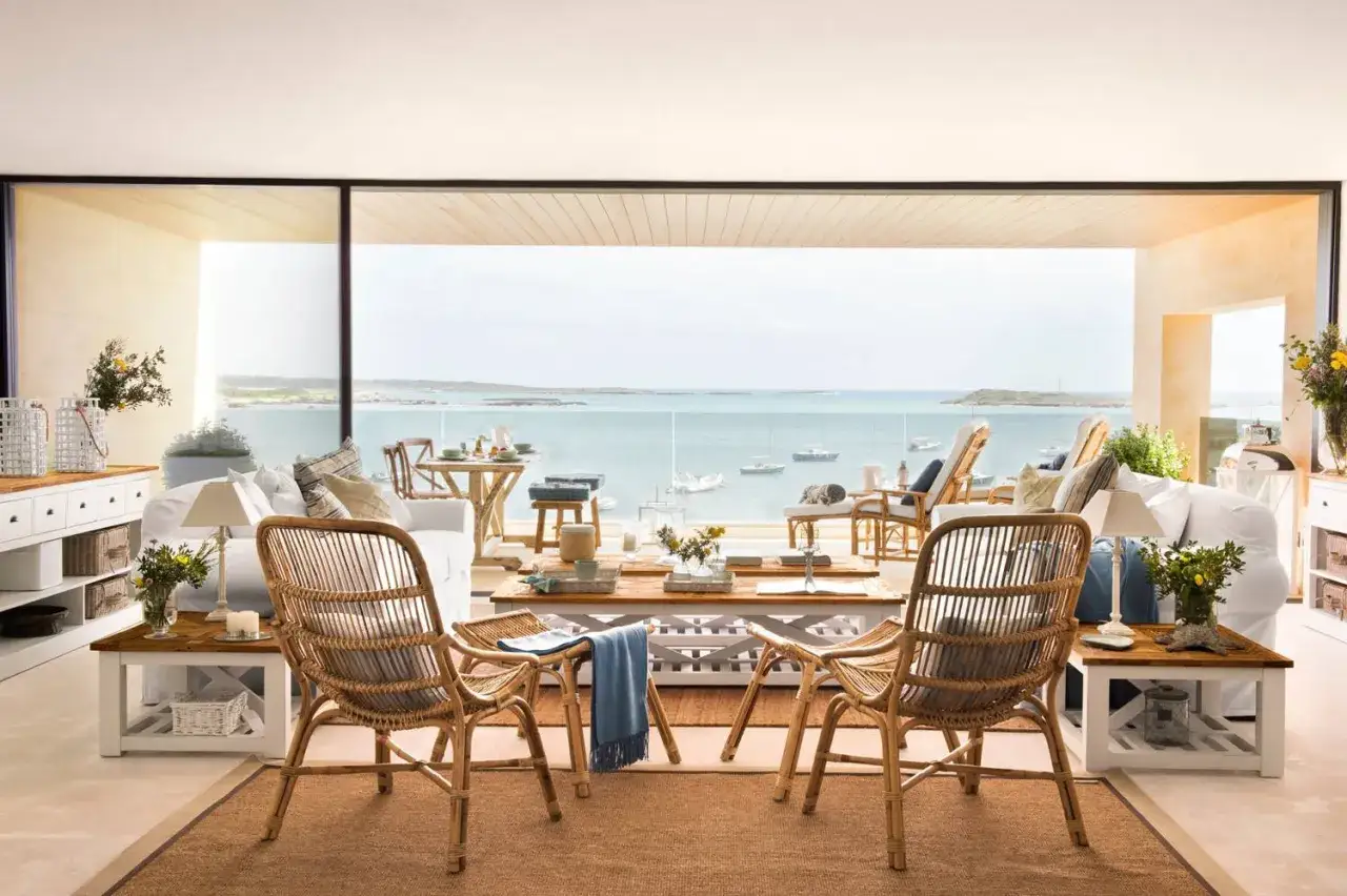 Salón con muebles de mimbre y sofás blancos, con una ventana panorámica que ofrece vistas al mar y barcos.