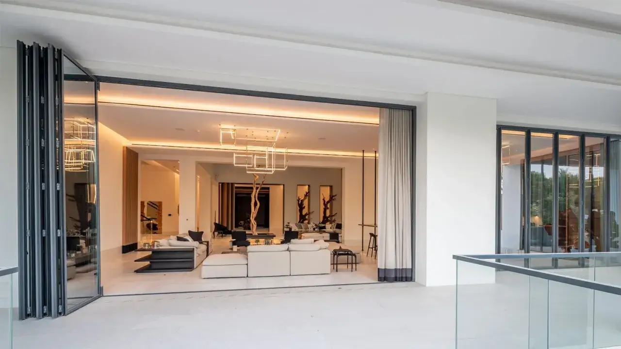 Amplio salón moderno con sofás blancos, una lámpara geométrica y una puerta abatible que conecta con el exterior.