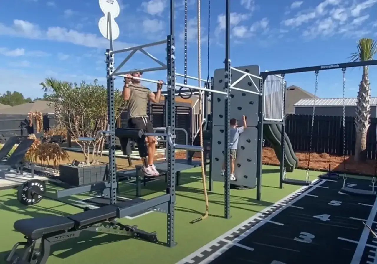 Un hombre hace dominadas en un gimnasio en el jard&iacute;n. Un ni&ntilde;o escala una pared. Hay equipo de entrenamiento y una pista de carreras.