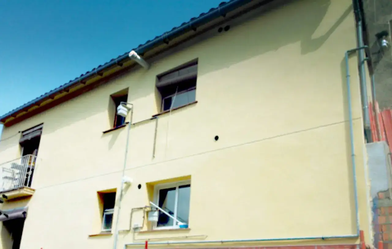 Fachada amarilla con ventanas y un balcón. Se aprecian tuberías y un tendedero, sugiriendo trabajos de mantenimiento o instalación. El mortero de cal se ve en buen estado.