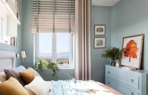 Acogedor plano dormitorio 9 metros cuadrados con cama, cómoda azul, cuadros y vistas a la ciudad.