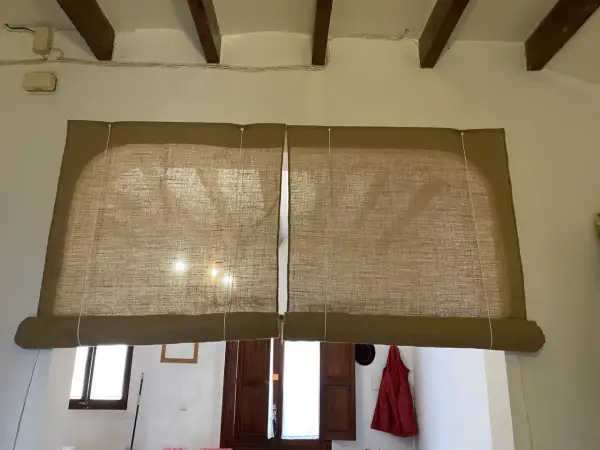 Dos estores enrollables caseros de tela de yute cubren una ventana. La luz del sol se filtra a través de ellos.
