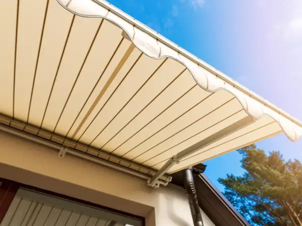 Toldo beige desplegado sobre terraza, cumpliendo la normativa. Cielo azul y árbol al fondo.
