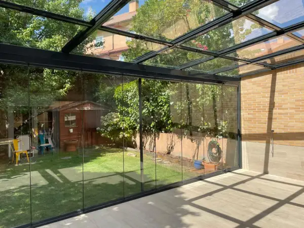 Modernos tejados para porches de cristal, con vistas a un jardín con caseta de madera y árboles frondosos.