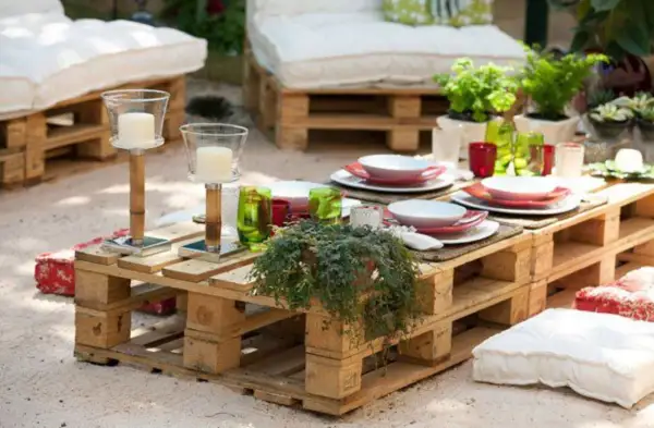 Mesa de jardín con ideas con palets, decorada con vajilla, velas y plantas. Cojines a los lados invitan a relajarse.