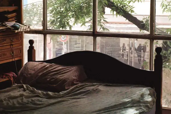 Dormitorio con ventana en el cabecero. La cama está hecha, con sábanas arrugadas y una almohada. A la izquierda, un mueble de madera con cajones.